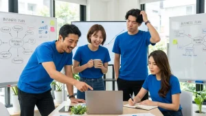 オフィスで青いTシャツを着た4人のビジネスチームが、ラップトップを囲んでビジネスプランを活発に議論している様子。笑顔で画面を指差す男性、頭をかいて困惑する男性、マグカップを持って笑う女性、真剣な表情でメモを取る女性が写っており、背景には戦略図が描かれたホワイトボードやグラフが表示されたモニターがある
